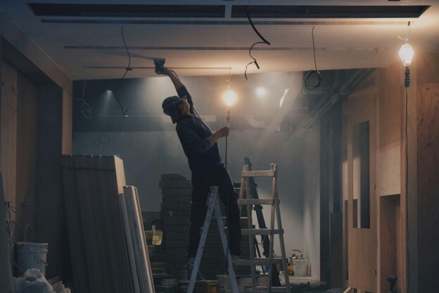 A worker stands on a ladder while holding a wire and bulb and working on the ceiling with a tool