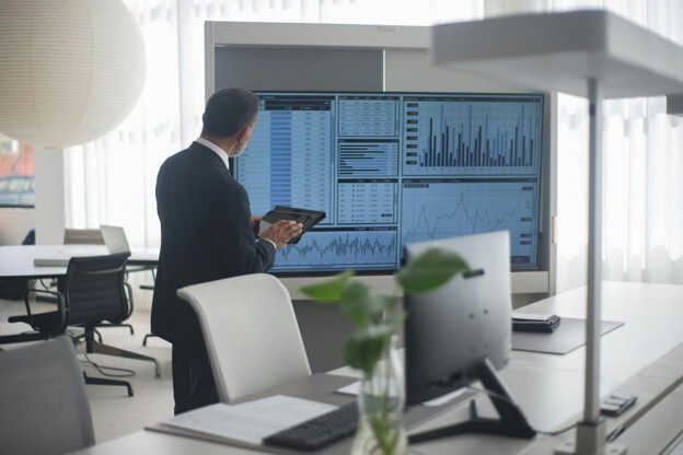 A man in a suit holds a tablet while looking at a large screen showing numbers and graphs in a meeting room