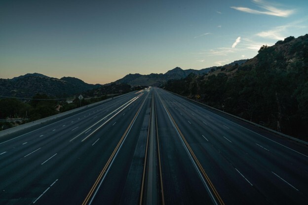 A smooth straight stretch of motorway with mountainous terrain on either side and a blue sky above
