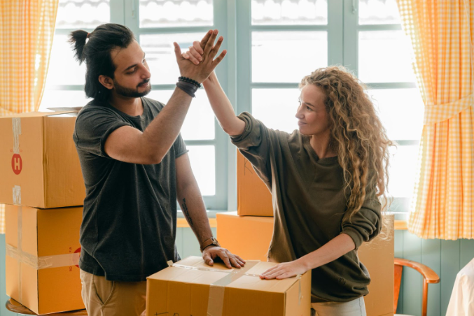 A man and a woman high-five over a taped carton with other boxes and a window visible behind them