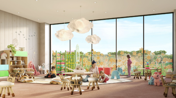 A large room with a pink carpet, children's furniture and toys has an adult sitting on the floor with a young toddler, while another child stands by a large window,w and one sits on the side