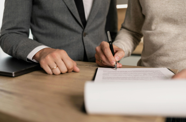 A person in a suit sits with another holding a pen above a document on a desk
