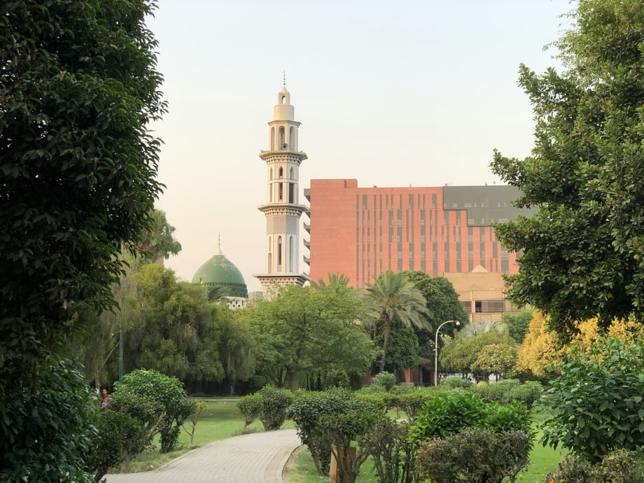 A path winding through foliage with a mosque and red brick building visible ahead
