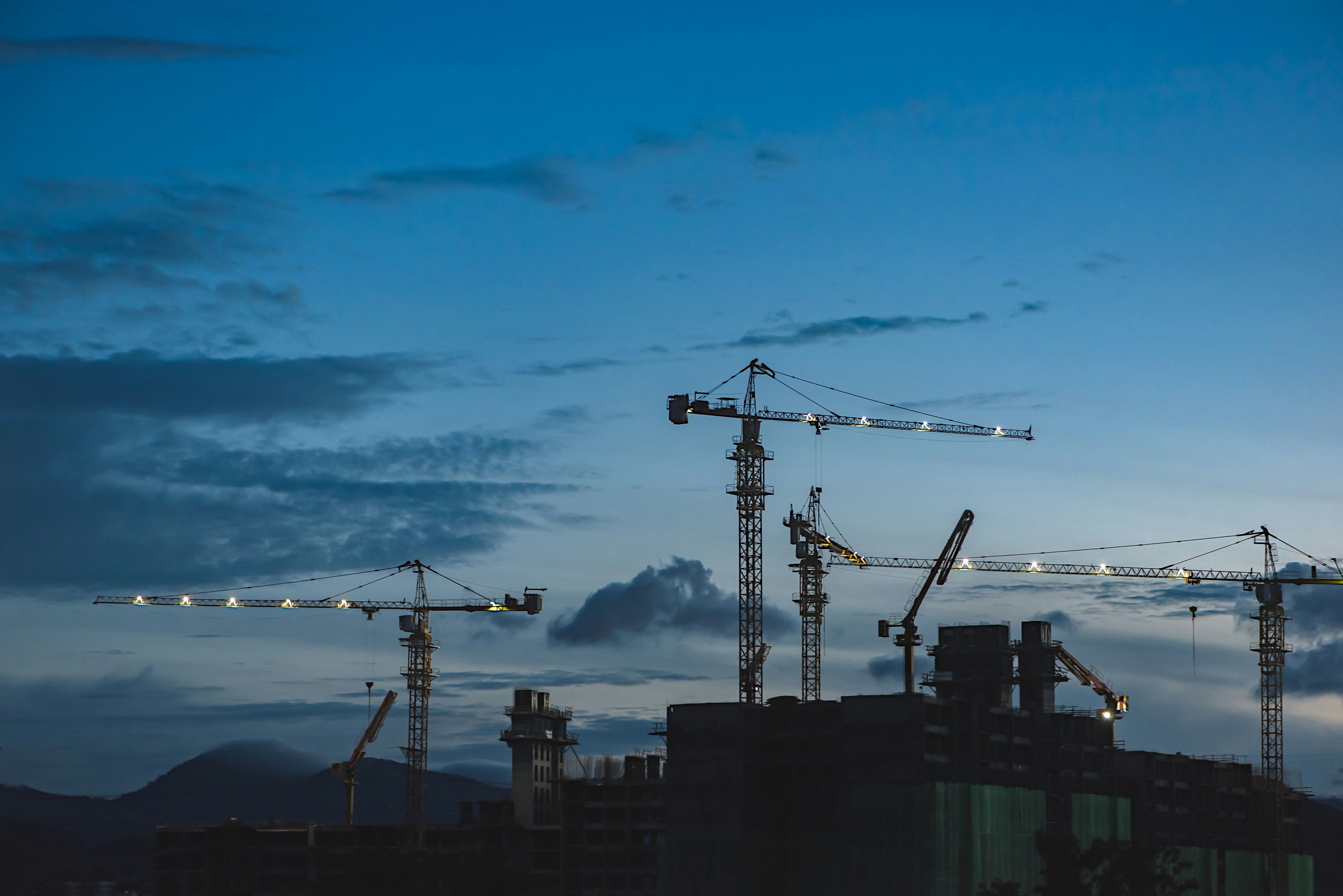 A silhouette of a building under construction with cranes around it against an evening sky and a backdrop of mountains