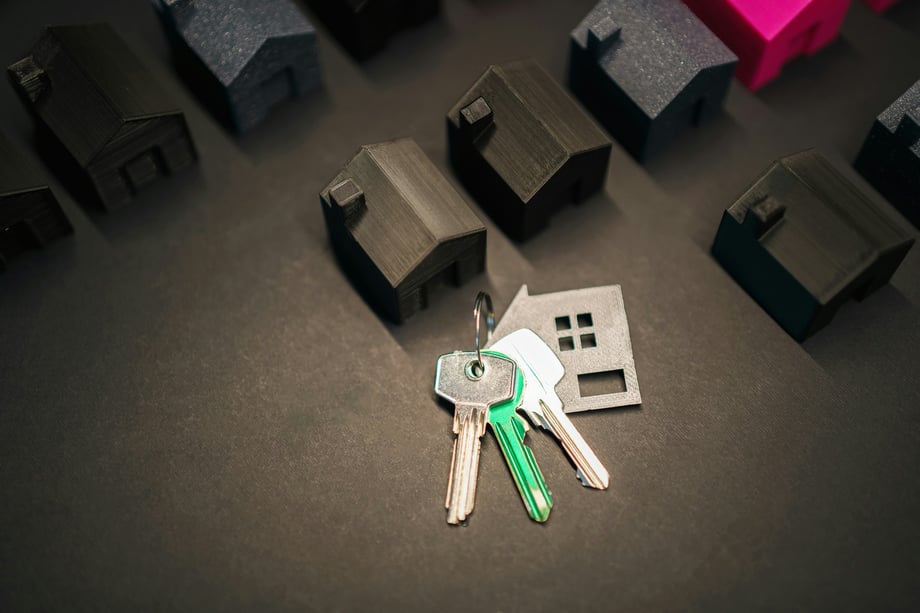 Small plastic houses in black, blue, and pink lined up on a black surface with a set of keys with a house cutout keychain