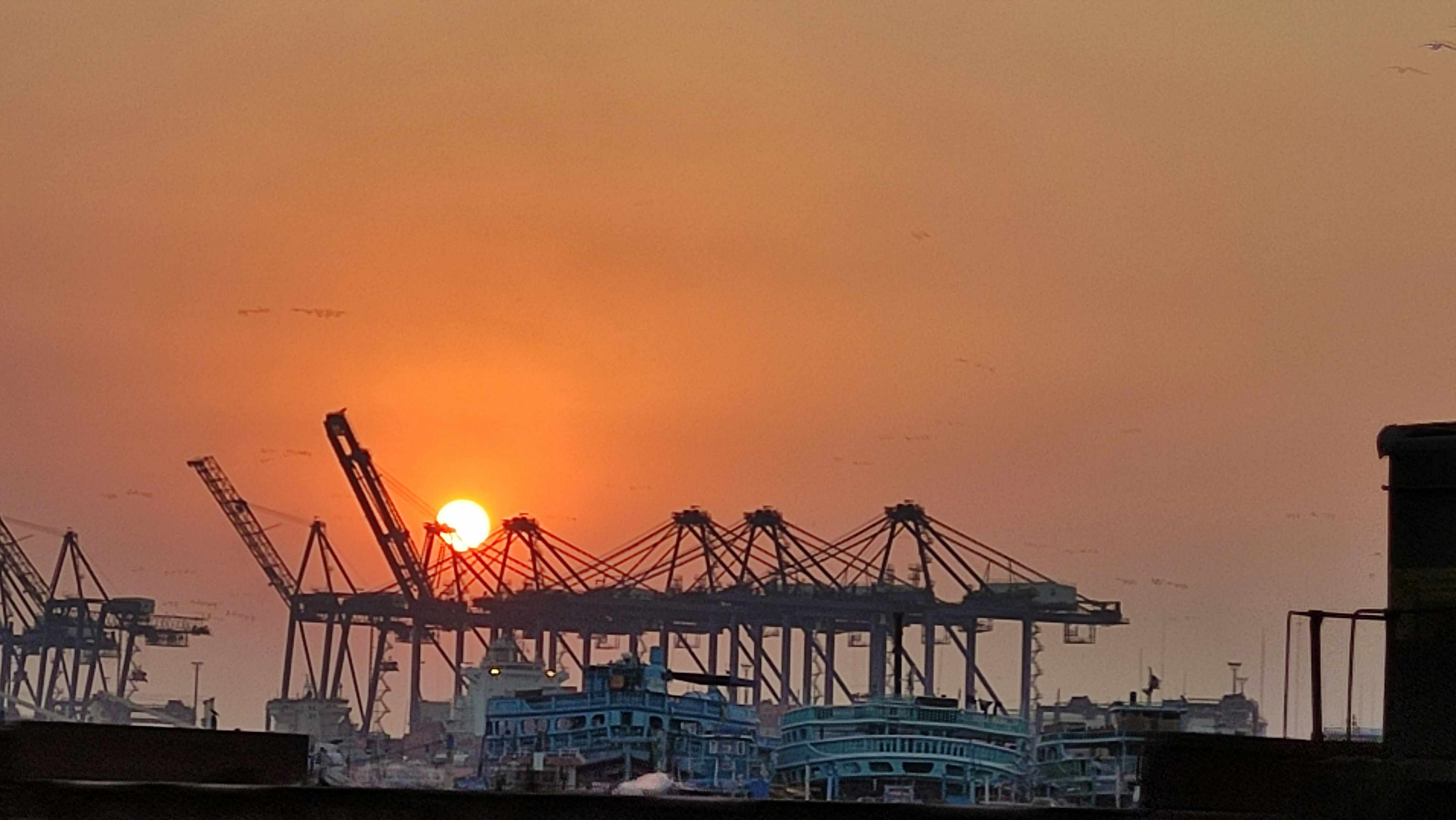 Boats docked in front of the silhouette of cranes at the Gwadar port as the sun shines low in the evening sky
