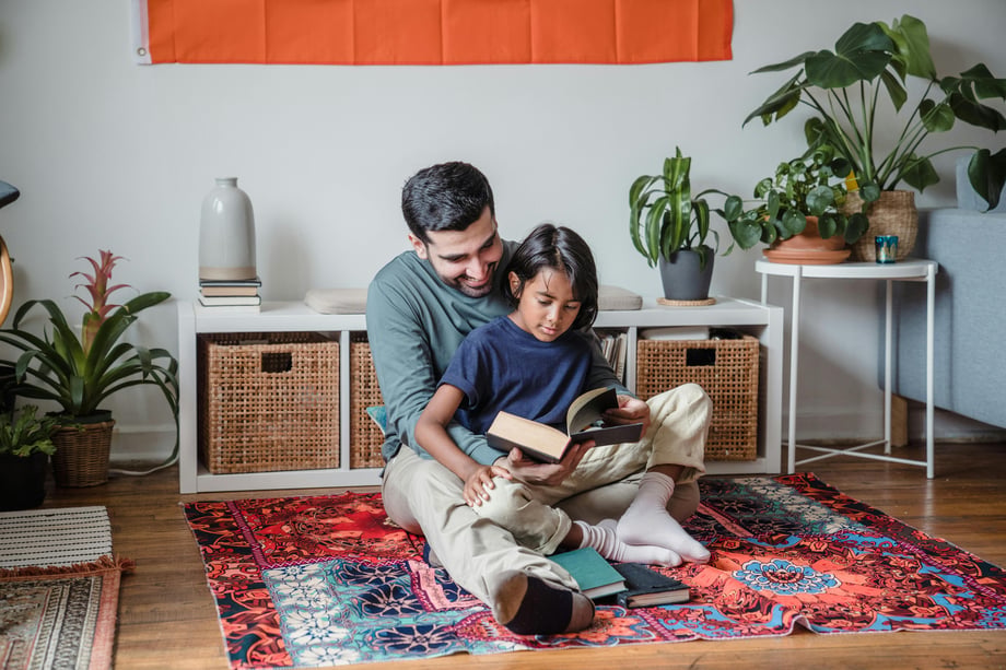  A Pakistani father and son sit together on a bright, colourful carpet with plants and storage in the background