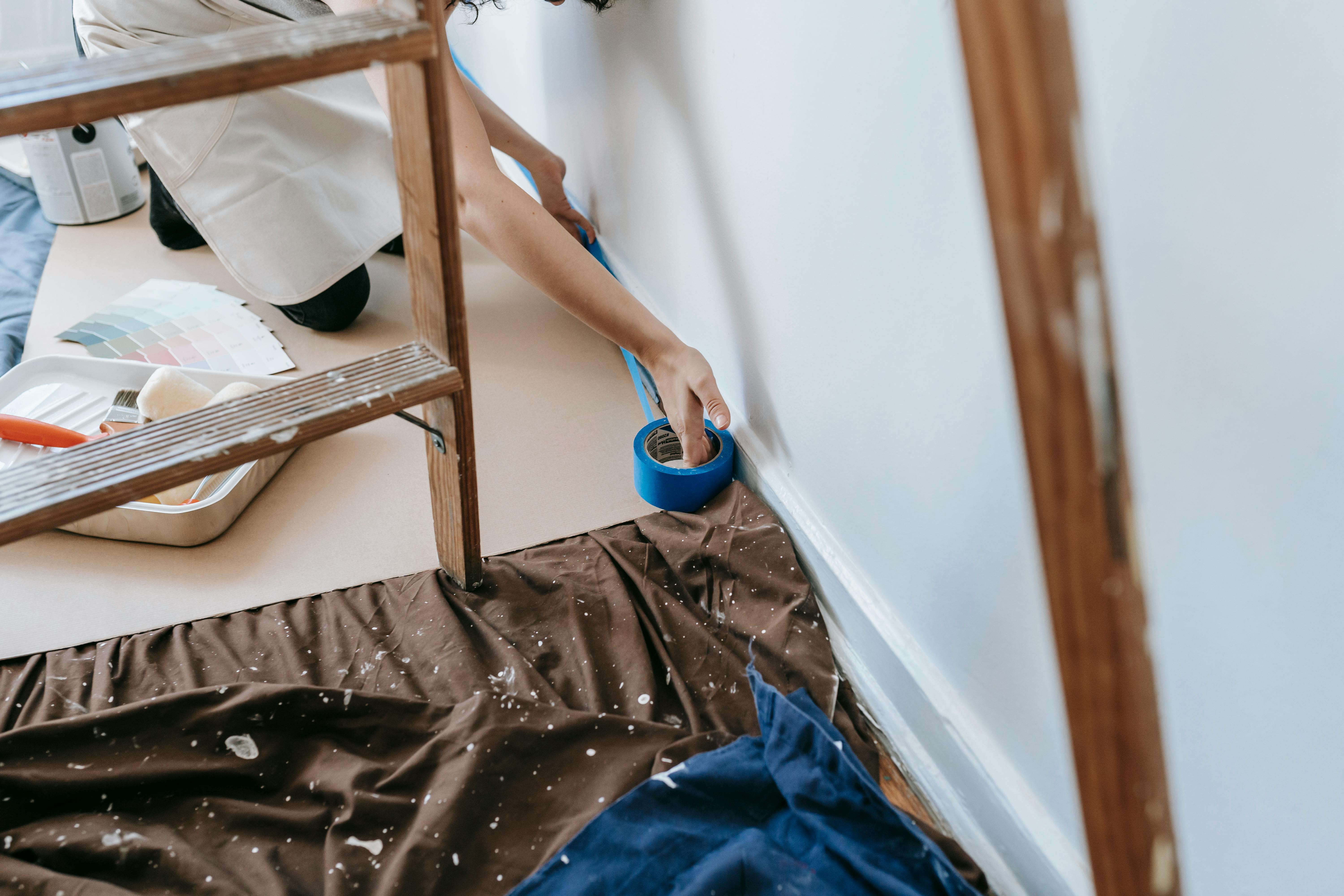 A person puts blue tape on the skirting with fabric spread out below a ladder as they get ready to paint