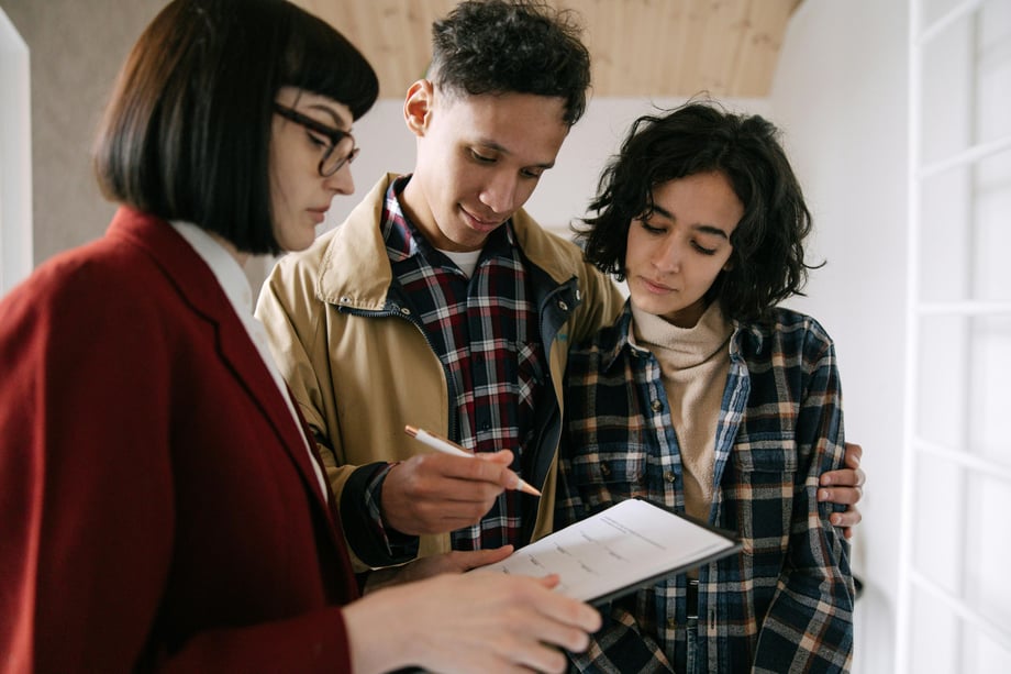 A couple in matching plaid shirts stand together looking over a form held by a woman in a wine red coat and glasses in a brightly lit indoor space