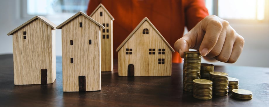 Wooden house figures of varyng shapes and sizes on a wooden table next to stacks of coins with a hand touching the tallest stack from behind the surface