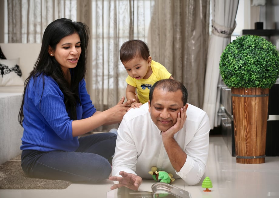 A man sits lies on his stomach on an apartment floor beside a woman with an infant on his back