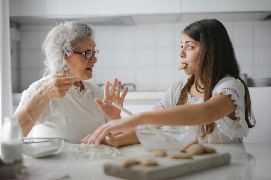 A younger female sits at a kitchen counter with a cookie in her mouth across from an older woman with silver hair