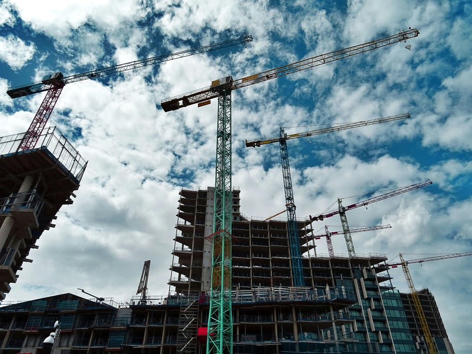 Six cranes seen towering over a luxury-building under construction against a blue sky, scattered with bright white clouds