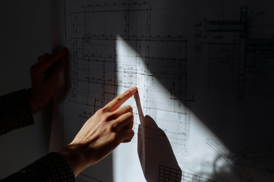  A blue print held against a wall while a hand points to a specific part on the drawing as sunlight streams onto the paper