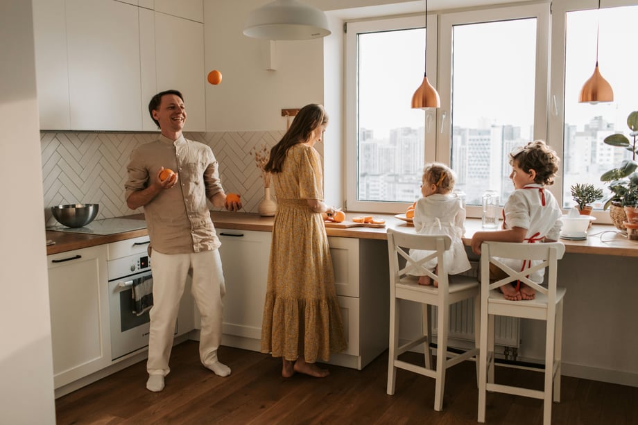 A man juggles oranges in a kitchen while a woman stands beside him cutting an orange and two children sit at bar chairs watching them