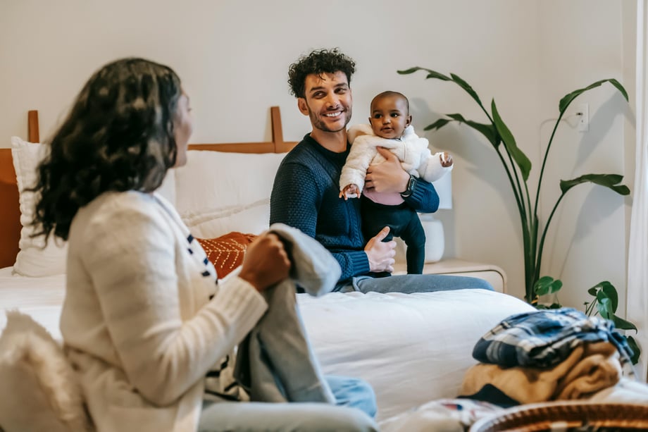 A man sits on a bed holding an infant while a woman sits across, folding clothes