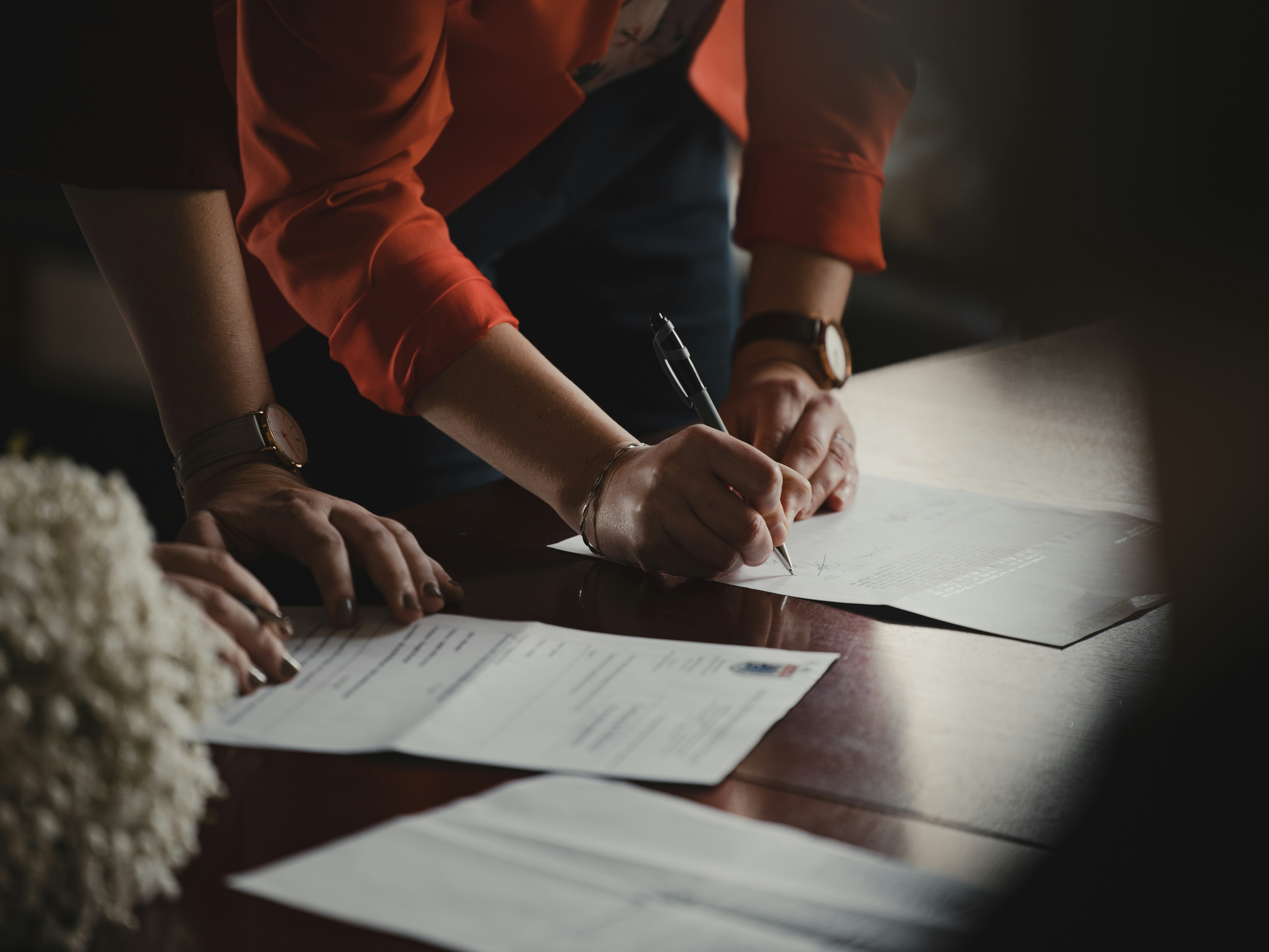 A person in orange holds a pen over a paper while another stands close by, both leaning over a table strewn with papers
