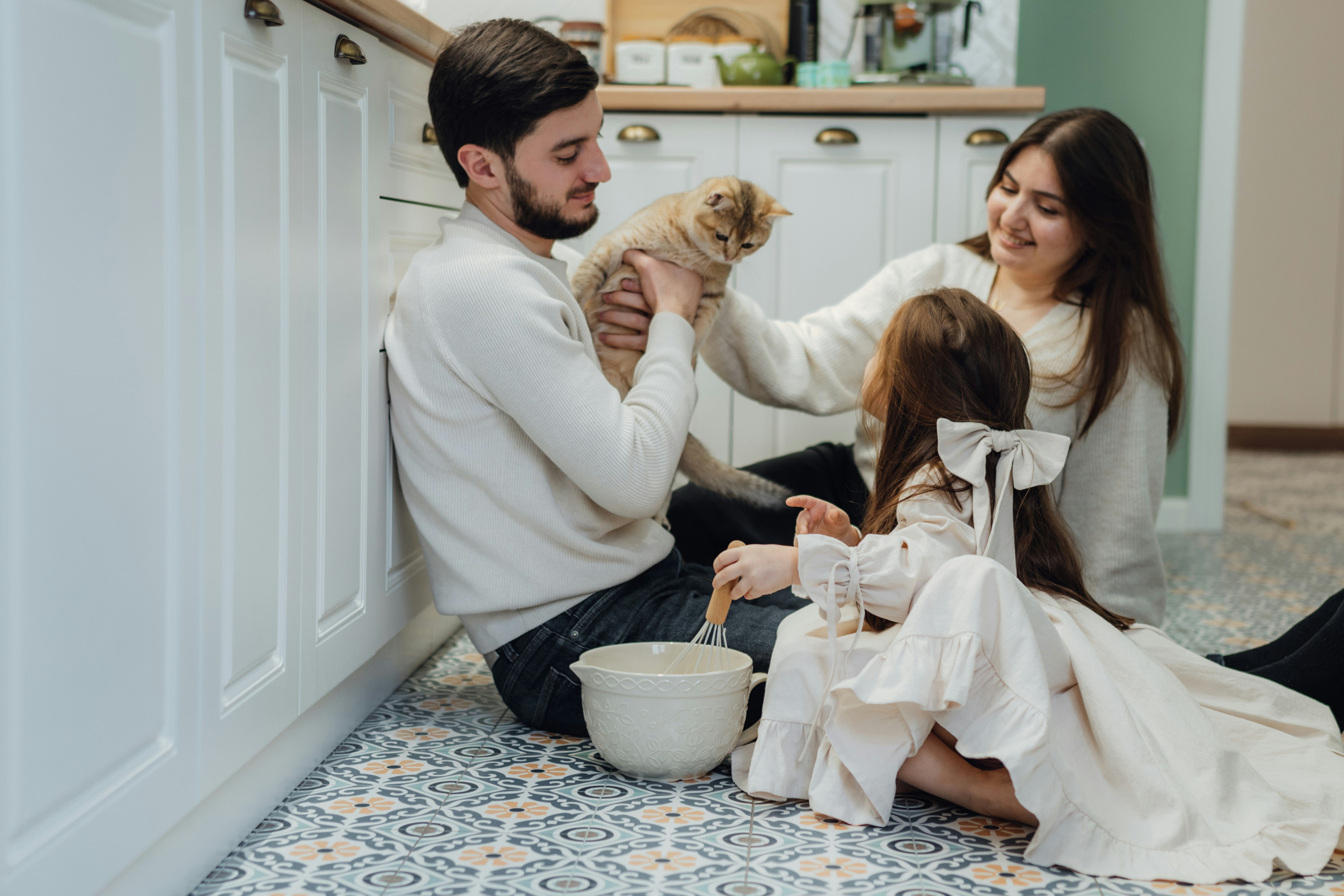 A man, a woman, and a child all dressed in white sit on a tiled kitchen floor while the man holds a cat