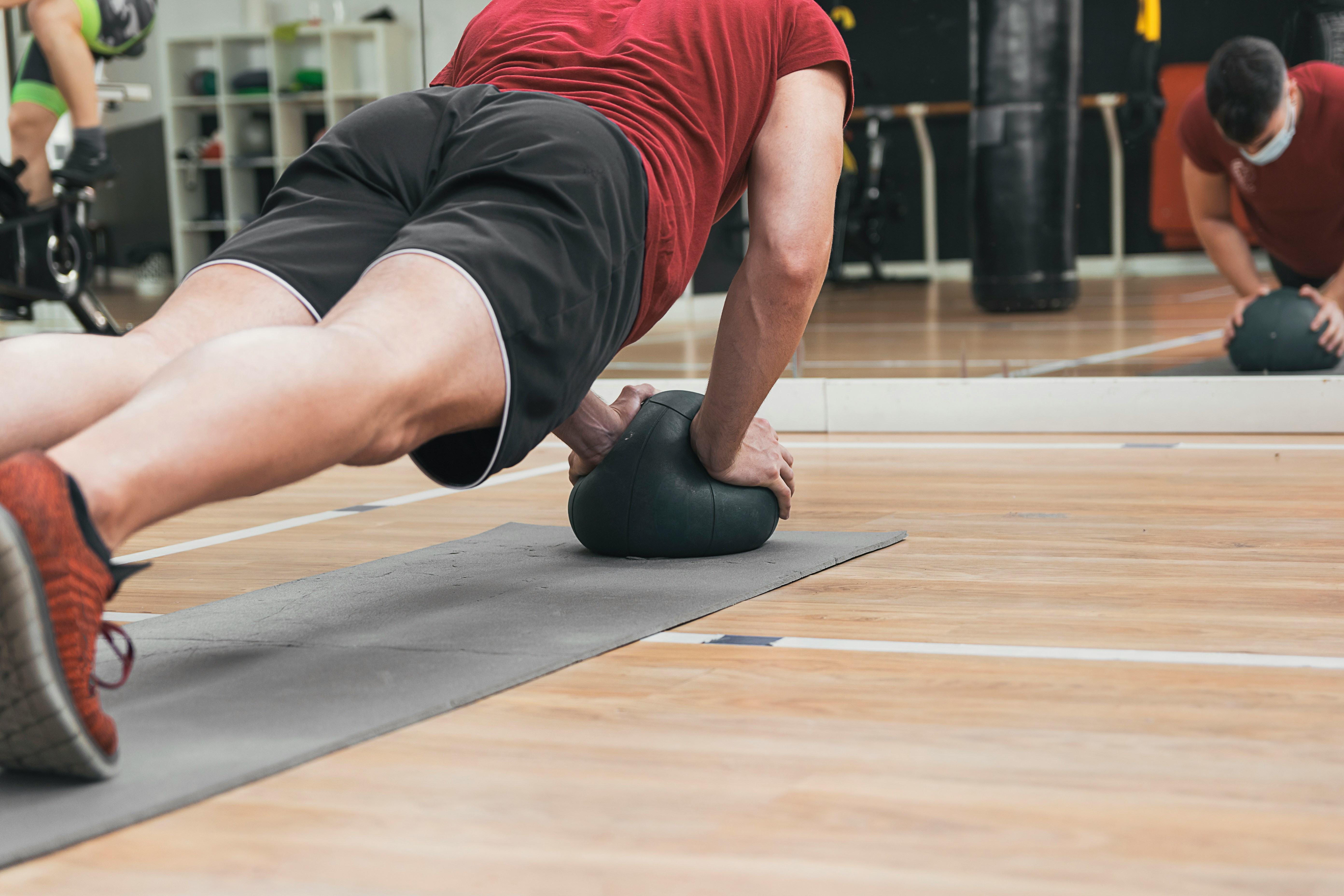 A person maintains a position on his toes with his hands resting on a medicine ball above a yoga mat, at a gym